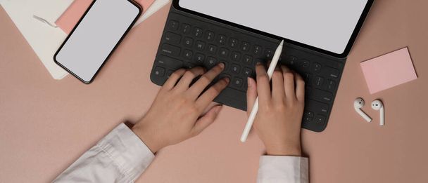 Fotografia aérea de mãos femininas digitando no teclado do tablet com smartphone, artigos de papelaria e acessórios na mesa rosa
 - Foto, Imagem