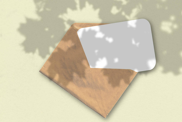 Een envelop met een vel getextureerd wit papier op de gele achtergrond. Mockup met een overlay van plantenschaduwen. Natuurlijk licht werpt schaduwen van een exotische plant. Horizontale oriëntatie. - Foto, afbeelding