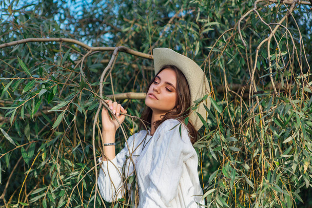 Young beautiful brunette woman dressed in a white sweater, jeans and cowboy straw hat standing close to the tree brunches with green leaves - Φωτογραφία, εικόνα