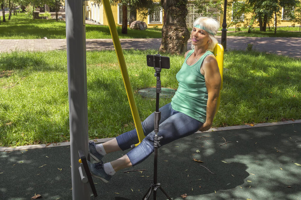 Mujer de edad avanzada delgada con el pelo gris lleva a cabo el entrenamiento virtual utilizando un teléfono móvil en un simulador en un parque de la ciudad durante las clases de fitness individuales
 - Foto, Imagen