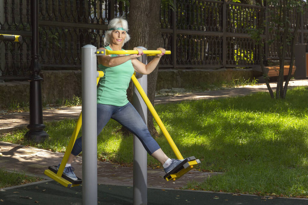 Mujer de edad avanzada delgada con el pelo gris conduce clases individuales de fitness en un simulador en un parque de la ciudad. Copiar espacio - Foto, imagen