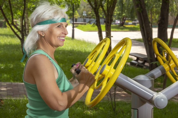 Mujer de edad avanzada delgada con el pelo gris conduce clases individuales de fitness en un simulador en un parque de la ciudad. Primer plano - Foto, imagen