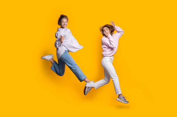 Zıplayan iki beyaz sarışın kadın sarı stüdyo duvarında oynayıp eğleniyorlar. - Fotoğraf, Görsel