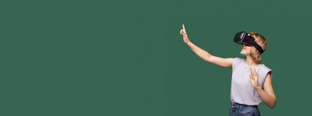 Bella giovane donna bionda con auricolare vr toccare qualcosa sullo spazio libero studio verde - Foto, immagini