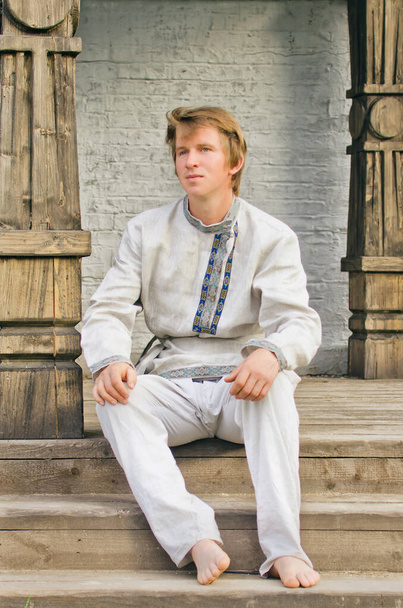 junger Mann in alten Leinenkleidern sitzt auf den Stufen der Veranda eines hölzernen Terems - Foto, Bild