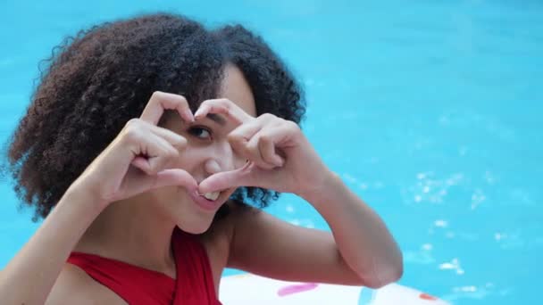 Joven estudiante africana rizada mujer en reposo sentada cerca de la piscina sonriendo amable mirando a la cámara haciendo corazón forma dedos doblando las manos en señal de amor y enviando un beso de aire a la cámara, de cerca retrato - Imágenes, Vídeo