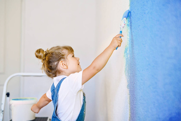 Pequeña niña divertida pintando la pared con color en una casa nueva. Casa de apartamento de reparación familiar. El niño feliz del bebé pinta la pared, eligiendo el color con la paleta, divirtiéndose con el cepillo, adentro - Foto, Imagen