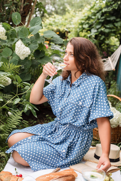 Zomer picknick. Een meisje in een zomerse blauwe jurk met stippen en een strohoed zit op een picknick in de natuur en drinkt mojito. Picknick met stokbrood, druiven, kersen en cocktail. - Foto, afbeelding