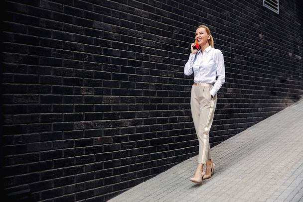 Pełna długość atrakcyjnej blondynki śmiejącej się modnej bizneswoman chodzącej po ulicy i rozmawiającej przez telefon. - Zdjęcie, obraz