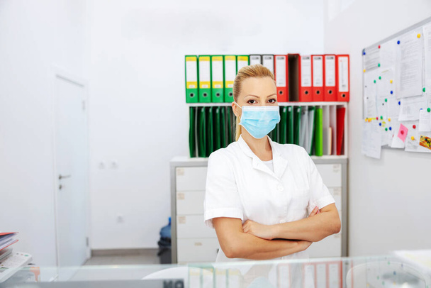 Attractive positive blond female lab assistant in sterile uniform with surgical mask on standing in laboratory with arms crossed and looking at camera. - Фото, изображение