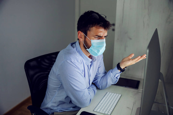 Young attractive bearded businessman with protective mask on sitting in office and trying to find solution for problem. - Photo, Image