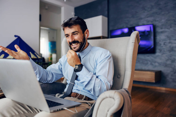 Junger schöner bärtiger Mann in eleganter Kleidung sitzt zu Hause im Stuhl und nutzt Laptop für Konferenzschaltung. - Foto, Bild