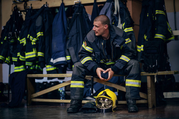 Jeune pompier attrayant en uniforme de protection assis dans la caserne de pompiers et attendant d'autres pompiers. Il est prêt pour l'action. - Photo, image