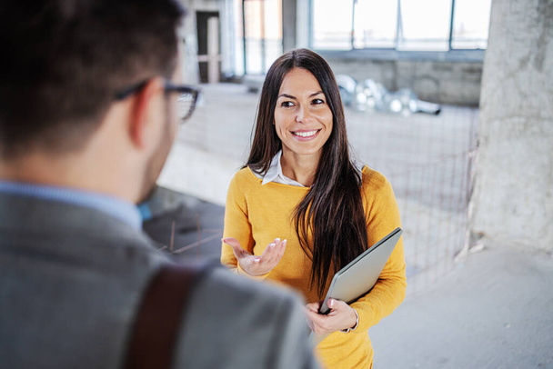 Mujer de negocios sonriente de pie en el edificio en proceso de construcción y hablando con su colega masculino. - Foto, imagen