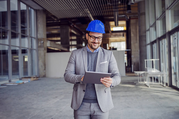 Portrait of attractive unshaven architect in suit with helmet on head walking around building in construction process and using tablet. - Photo, Image