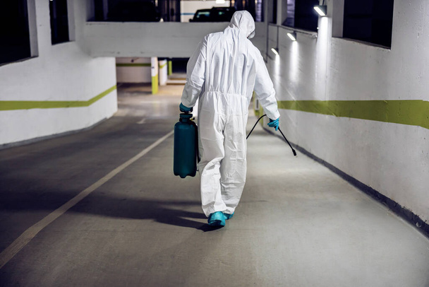 Εργαζόμενος σε αποστειρωμένη στολή και μάσκα περπάτημα γούρνα υπόγειο γκαράζ και την πόρτα της επιφάνειας. Προστασία από τον ιό του στέμματος / έννοια covid-19. - Φωτογραφία, εικόνα