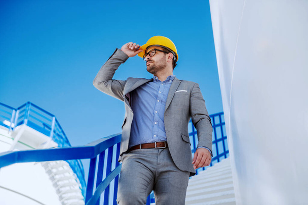 Young rich attractive caucasian businessman in suit with helmet on head going down the stairs, holding helmet and looking around. Refinery exterior. - Valokuva, kuva