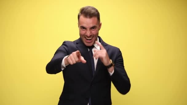 entuzjastyczny elegancki mężczyzna w garniturze wytykający palcami, śmiejący się, robiący kciuki w górę gest, tańczący, poruszający się, trzymający pięści w powietrzu i świętujący zwycięstwo na żółtym tle - Materiał filmowy, wideo