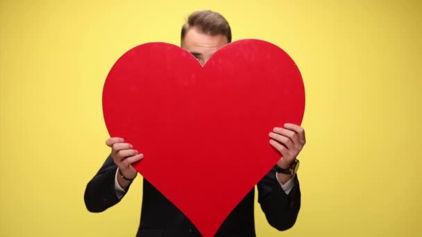 unhappy young businessman covering face with big red heart, revealing himself, embracing the heart and looking sadly down on yellow background - Footage, Video