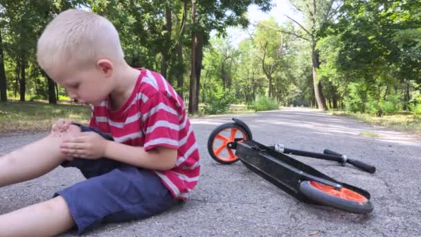 Un niño de cuatro años se cayó de una moto y se lesionó la rodilla. El niño llora, se frota la rodilla y sopla sobre ella. El concepto de conducción descuidada y descuido parental. - Metraje, vídeo
