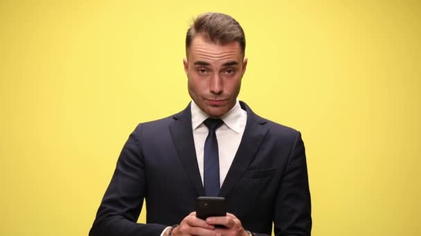 sexy joven hombre de negocios en traje sosteniendo teléfono escribir un mensaje mirando hacia arriba y sonriendo sobre fondo amarillo - Imágenes, Vídeo