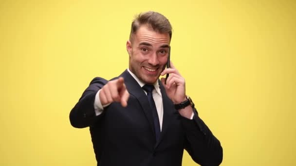 smiling young man in suit talking on the phone pointing fingers and making call me gesture on yellow background - Footage, Video