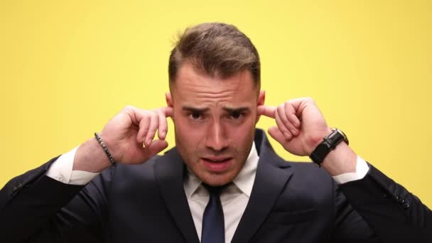elegáns fiatal üzletember öltönyben borító fülek ujjaival megállítja a zajt, vicces arcok és mosolygós sárga háttér - Felvétel, videó