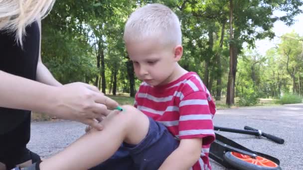 Een kind van vier viel van een scooter en verwondde zijn knie. Moeder steekt de wond op haar knie met briljant groen en brengt een pleister. - Video