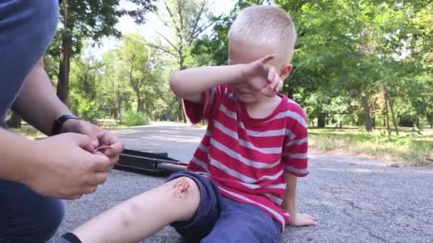 Ein vierjähriger Junge stürzte von einem Roller und brach sich das Knie. Papa leistet Erste Hilfe, indem er die Wunde desinfiziert und ein Pflaster aufträgt. - Filmmaterial, Video