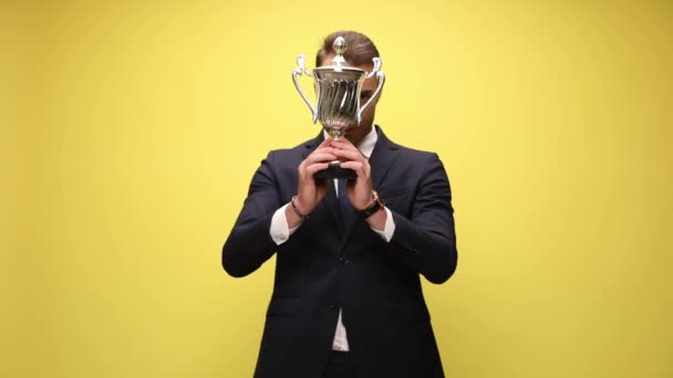głupi młody biznesmen chowający się za srebrnym trofeum, patrząc na trofeum i ujawniając się, uśmiechając się i stojąc na żółtym tle - Materiał filmowy, wideo