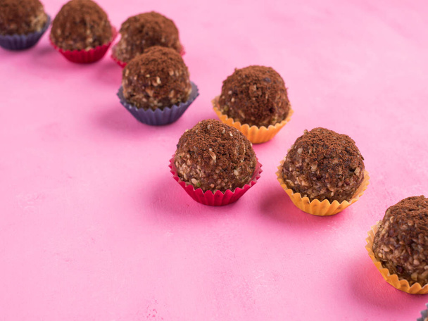 Home made vegan energy protein balls with oats, nuts, almond butter, dates, dried fruit, flax seeds, chocolate nibs in colorful paper cases on pink wooden background. Closeup - Фото, изображение