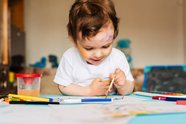 Fröhliches fröhliches Kind zeichnet mit Pinsel in Album mit vielen Malwerkzeugen. - Foto, Bild