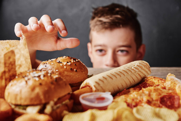 Rapaz faminto olha para comida saborosa e tomar hambúrguer da mesa. Conceito alimentar pouco saudável - Foto, Imagem