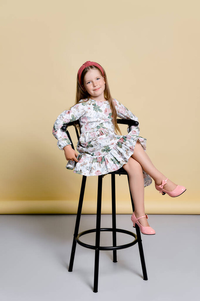 Cute little girl in sundress and hair band sits on tall chair in studio over pale yellow background - Photo, image