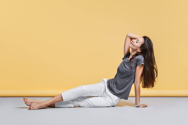Happy barefoot young woman in casual trousers and shirt sits on the floor in studio on yellow background - Foto, Imagen