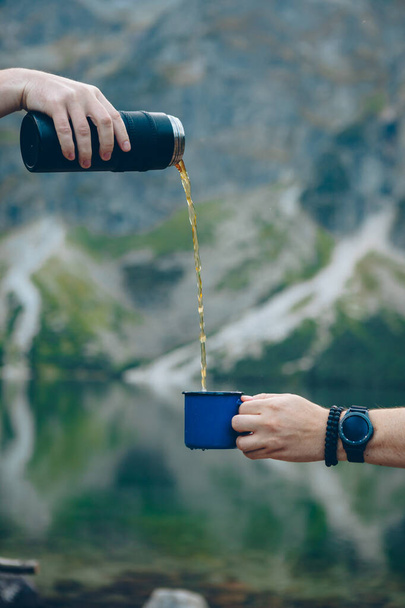 pouring tea from thermos to cup hiking concept lake with mountains on background warm up drink - Fotó, kép