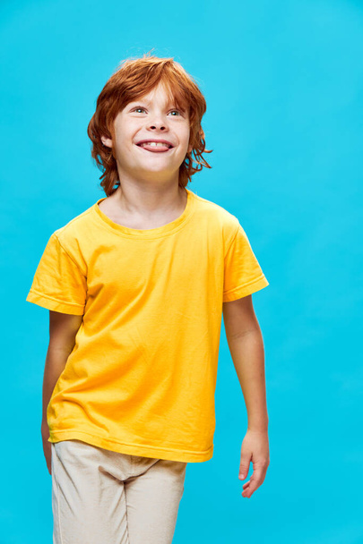 Happy red hair child in a T-shirt and trousers on a blue background shows his tongue - Photo, Image