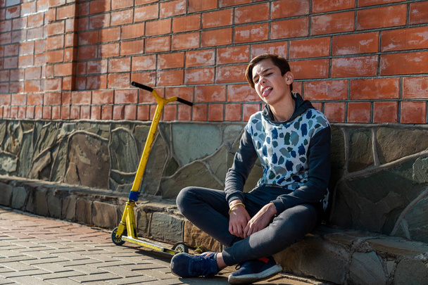 Um adolescente senta-se na calçada ao lado de uma scooter, e destaca a língua. Parede de tijolo no fundo. O conceito de adolescência e emoções
. - Foto, Imagem