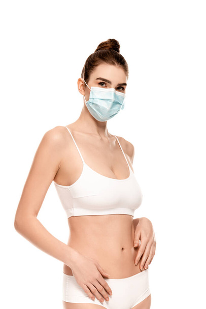 mladá žena v lékařské masce, horní a kalhotky při pohledu na kameru izolované na bílém - Fotografie, Obrázek