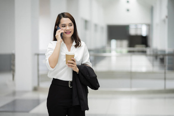 Retrato de uma jovem mulher de negócios asiática falando por telefone celular e segurando uma xícara de café no prédio de negócios. Foto de menina bonita em suite casual com telefone e xícara de café. Foto stock
 - Foto, Imagem