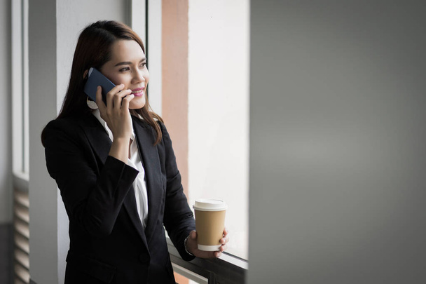 Portrait of a young Asian business woman talking over cellphone and holding cup of coffee in business building. Photo of beautiful girl in casual suite with phone and cup of coffee. Stock photo - Foto, afbeelding