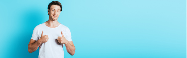 Горизонтальне зображення чоловіка в білій футболці, що показує великі пальці вгору на синьому фоні
 - Фото, зображення