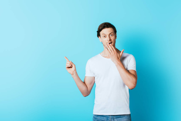 Uomo eccitato in t-shirt bianca che punta con il dito su sfondo blu - Foto, immagini