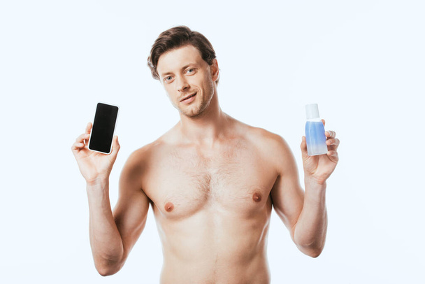 Muscular man holding bottle of lotion and smartphone isolated on white - Photo, Image