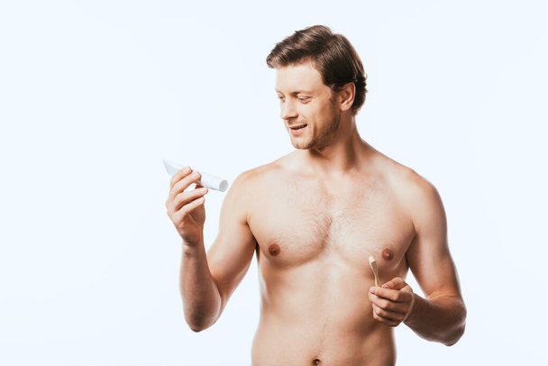 Shirtless man holding toothbrush while looking at toothpaste isolated on white - Foto, afbeelding