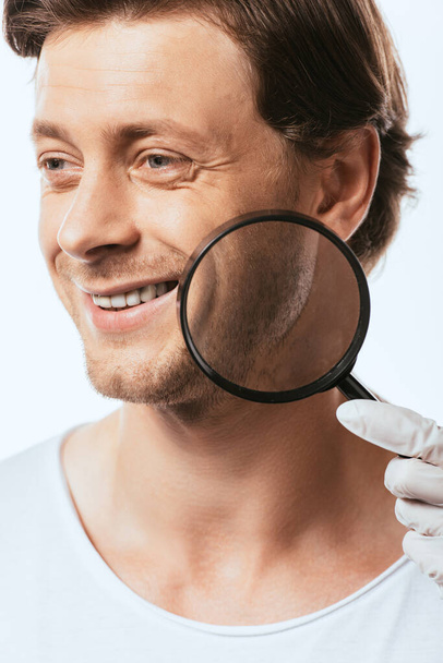 Dermatologist holding magnifying glass near cheek of young man isolated on white - Foto, immagini