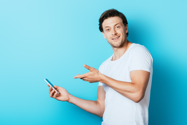 Beyaz tişörtlü genç adam mavi arka plandaki akıllı telefonu işaret ediyor. - Fotoğraf, Görsel