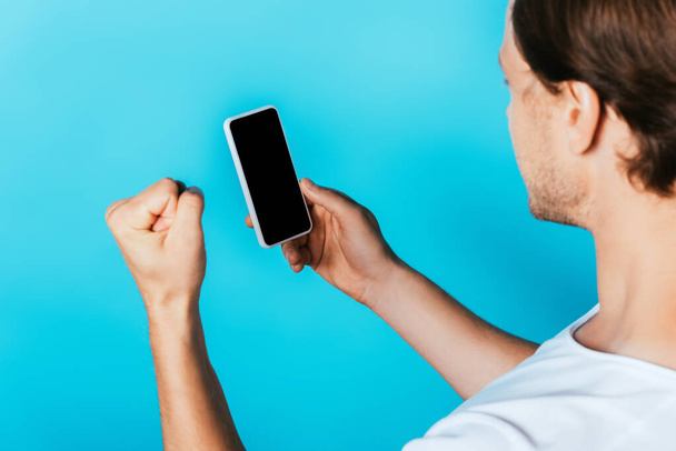 Man showing yeah gesture and holding smartphone with blank screen on blue background - Photo, Image