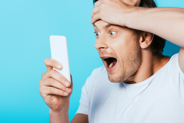 Uomo scioccato con mano vicino alla testa guardando smartphone su sfondo blu - Foto, immagini