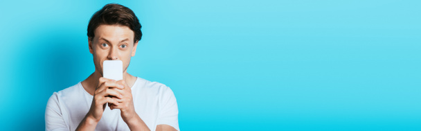 Panoramic shot of young man looking at camera while holding smartphone near mouth on blue background - Photo, Image